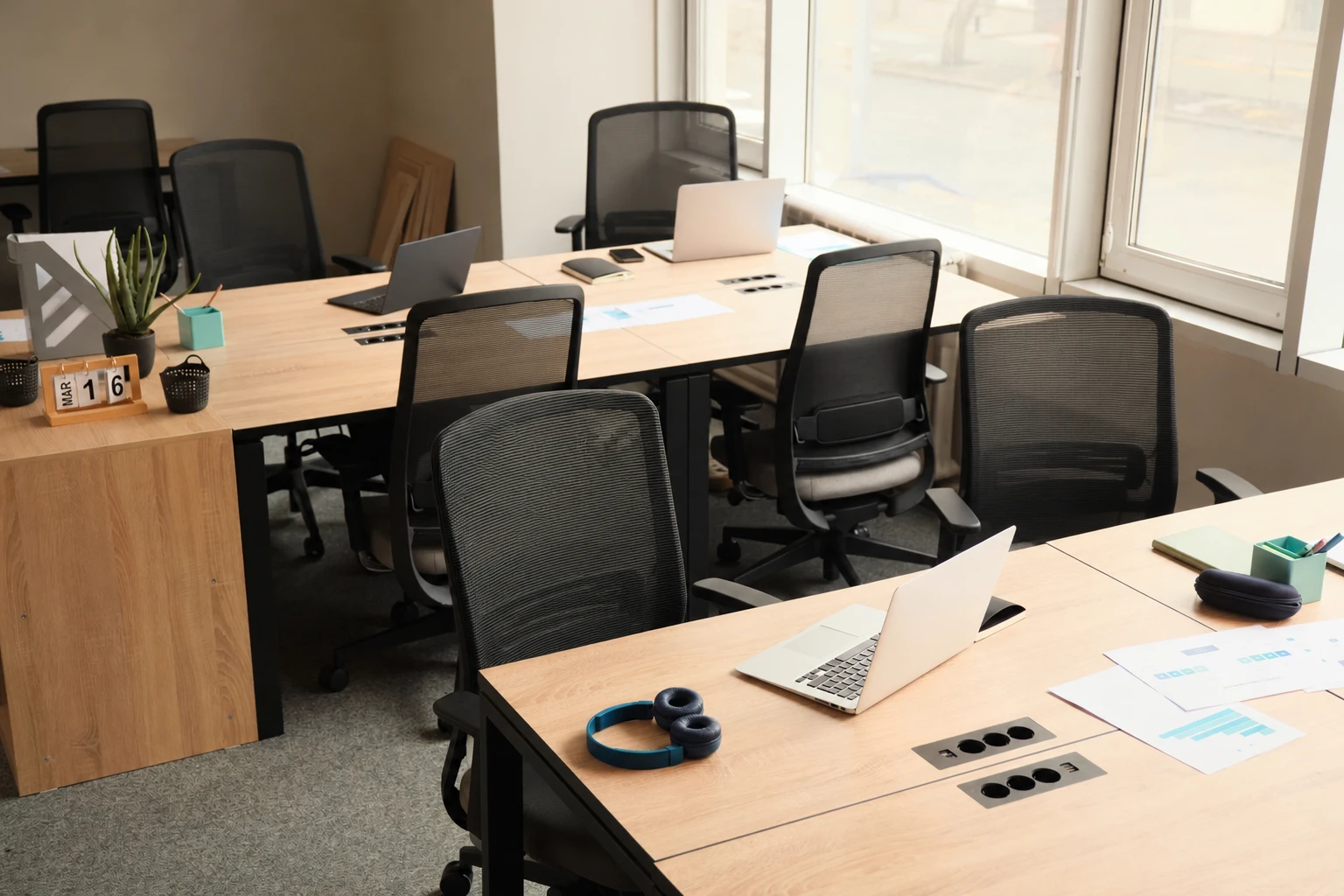 A sunlit shared office with wooden desks, laptops, and empty chairs.